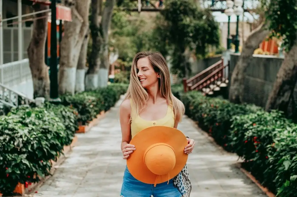 Smiling woman holding an orange sun hat while walking along a sunny tree-lined garden path.